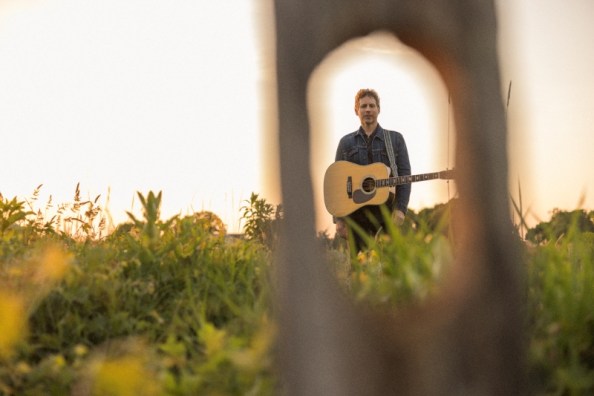 Jesse Terry stands in a field, with a guitar hanging in front of him from a guitar strap over his shoulder