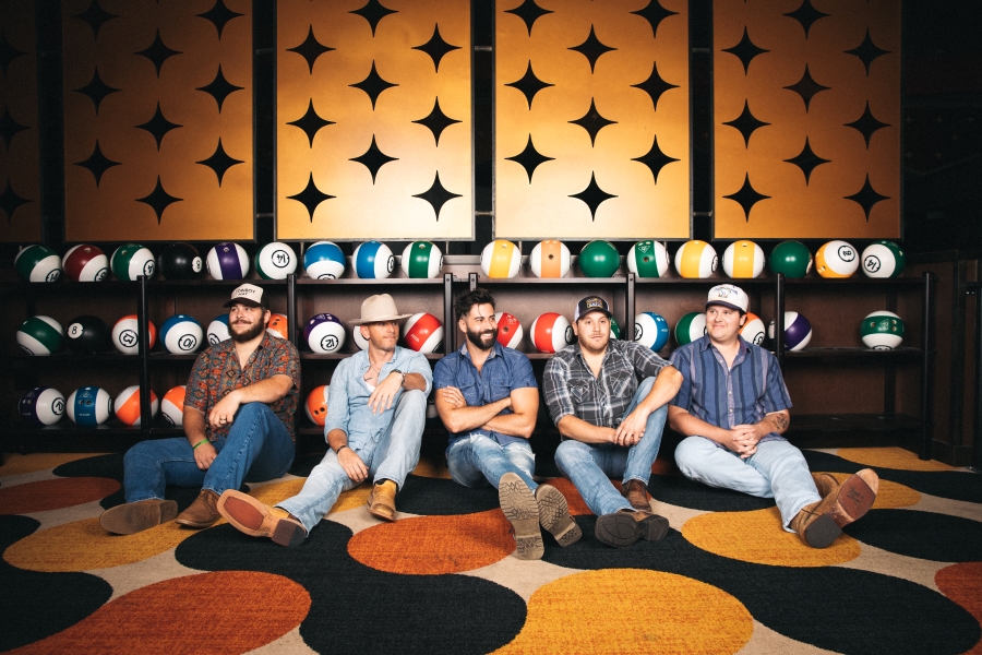 Midnight South band photo: four men sit on carpeting in a bowling alley in front of a row of bowling ball racks
